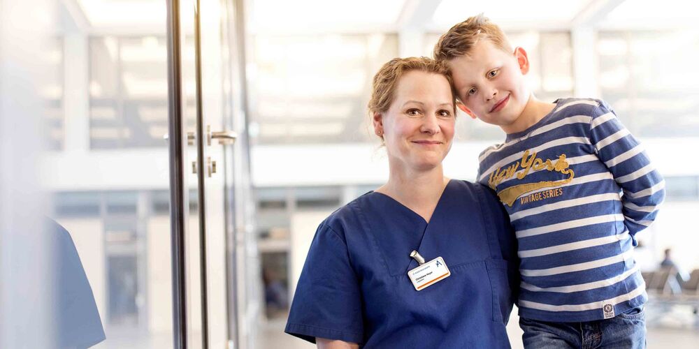 Eine Frau in Krankenhauskleidung kniet neben einem kleinen Jungen, welcher sich mit dem Kopf und Oberkörper an diese lehnt. Beide schauen lächelnd in die Kamera.