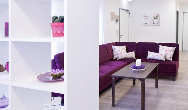 Zimmer der Behindertenhilfe. In diesem steht ein großes Sofa mit einem Couchtisch davor. Außerdem befindet sich ein offenes Regal als Raumtrenner in diesem und einem geschlossenem Regal an der Wand. 