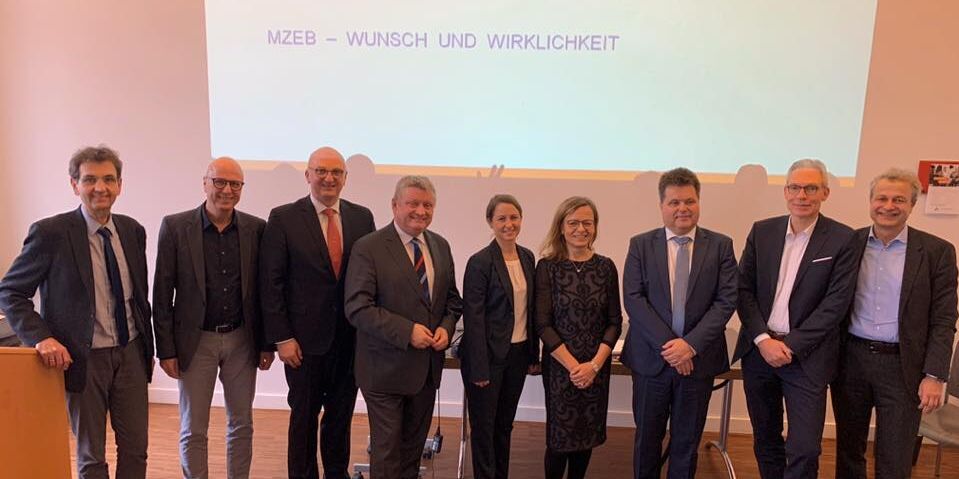Einige Herren und zwei Damen auf einer Fläche vor einer Präsentation, welche an die Wand geworfen wird.