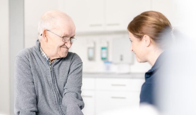 Ein älterer Patient sitzt lächelnd auf einem Patientenbett und schaut eine junge Mitarbeiterin an. 