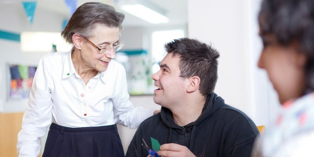 Eine ältere Dame steht neben einem jungen Mann, welcher an einem Tisch sitzt und bastelt. Sie hat ihm eine Hand auf die Schulter gelegt. Eine weitere jüngere Frau sitzt mit am Tisch. 