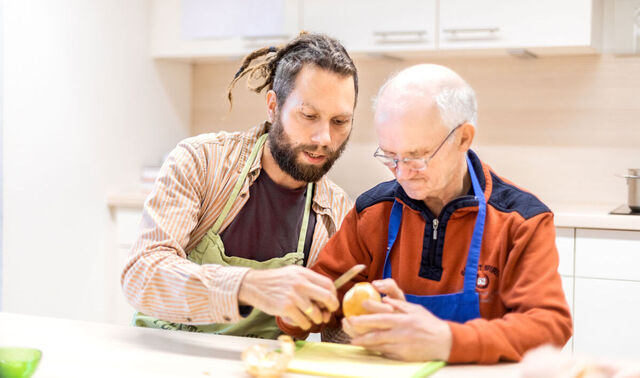 Ein älterer Mann und ein junger Pfleger sitzen gemeinsam in der Küche an einem Tisch. Der ältere Mann schält eine Kartoffel und kriegt hierbei Hilfe von dem jüngeren.