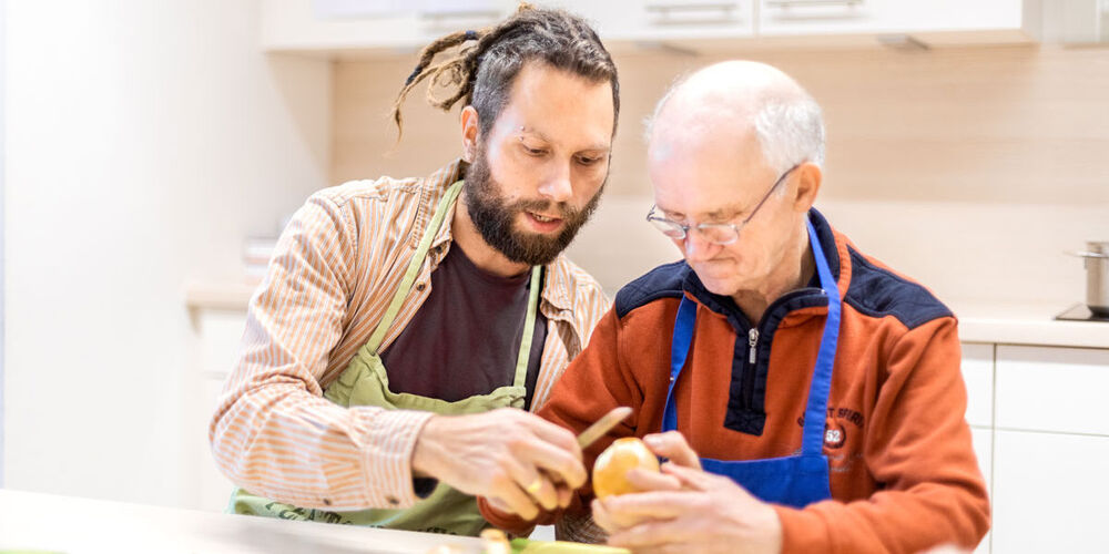 Ein älterer Mann und ein junger Pfleger sitzen gemeinsam in der Küche an einem Tisch. Der ältere Mann schält eine Kartoffel und kriegt hierbei Hilfe von dem jüngeren.
